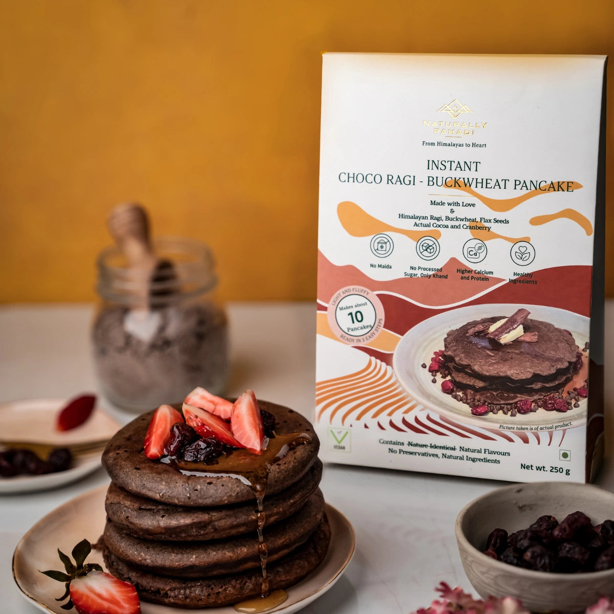 Stack of chocolate pancakes with berries on a plate next to an Instant Choco Ragi Buckwheat Pancake box.